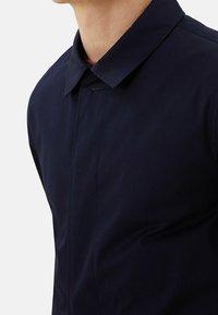 Hombre vistiendo una chaqueta de color azul marino oscuro con cuello, con una lista de botones oculta y una camisa roja debajo.