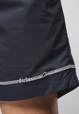 Granatowe szorty z białym haftowanym napisem i potrójnym paskiem przy brzegu na nodze osoby na jednolitym tle.