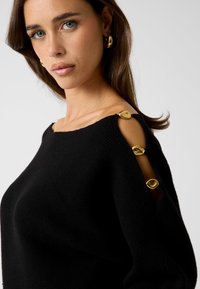 Zwarte gebreide trui met een off-shoulder ontwerp, voorzien van gouden knoppenaccenten op de mouwen en een gestructureerde afwerking.