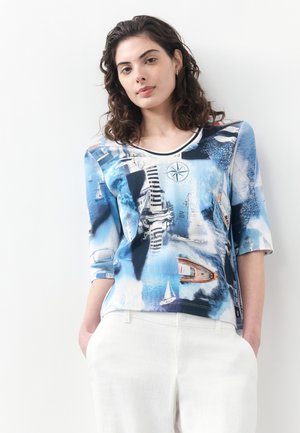 Frau mit lockigem Haar, die ein blaues T-Shirt mit nautischem Motiv und weiße Hosen trägt, mit den Händen in den Taschen stehend vor weißem Hintergrund.