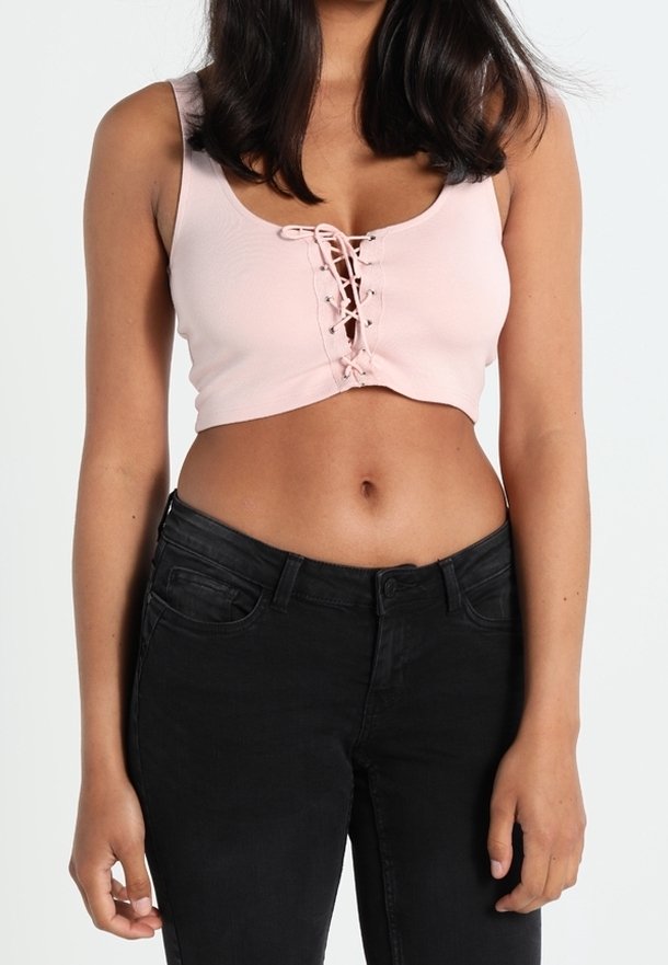 Femme portant un crop top sans manches rose clair avec un laçage avant et un jean noir taille haute, debout devant un fond blanc.
