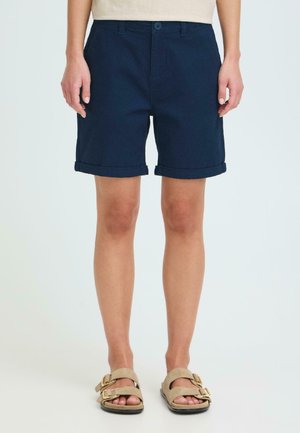 Persoon draagt marineblauwe shorts met omgeslagen zoom, beige sandalen met gouden gespen, staand tegen een effen lichte achtergrond.