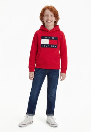 Enfant roux souriant portant un sweat à capuche rouge Tommy Hilfiger, un jean bleu et des baskets blanches, debout devant un fond blanc.