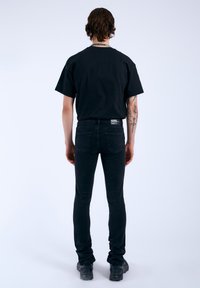 Camiseta de algodón negra de manga corta combinada con jeans de mezclilla oscuros. El modelo está de espaldas, mostrando una silueta ajustada y detalles mínimos.
