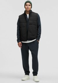 Chaleco acolchado negro con diseño de rombos, sobre una sudadera azul marino. Combinado con pantalones de chándal oscuros y zapatillas blancas, rematado con un gorro verde.