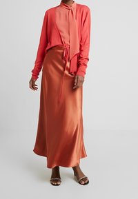 Cardigan en corail sur une blouse en satin assortie avec un nœud, associé à une longue jupe en satin fluide orange. Sandales à talons noires avec des détails.
