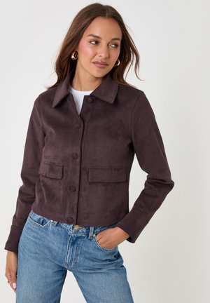 Mujer con chaqueta marrón oscuro con botones delanteros y bolsillos con solapa y vaqueros azules sobre un fondo blanco liso.