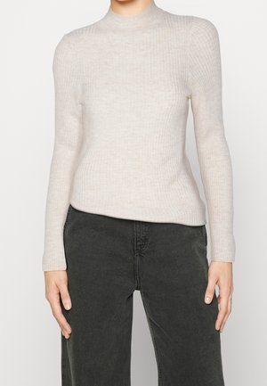 Person trägt einen cremefarbenen, gerippten Langarm-Pullover mit Stehkragen und schwarze, hoch taillierte Jeans, steht vor einem schlichten Hintergrund.