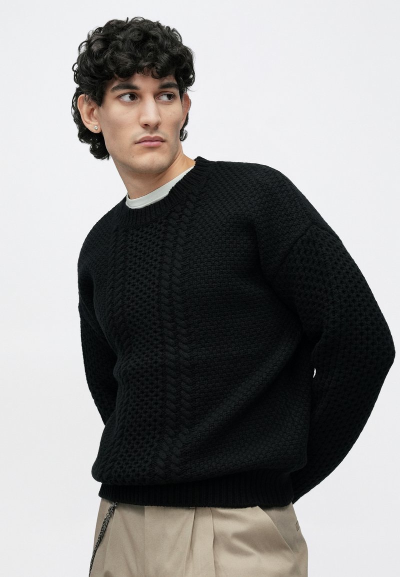 Young man with curly hair wearing a black textured knit sweater and beige pants, looking to the side against a plain background.