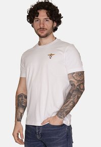 T-shirt bianca in cotone con maniche corte, collo rotondo e logo ricamato dorato e blu marino sul petto. Indossata con jeans blu scuro.