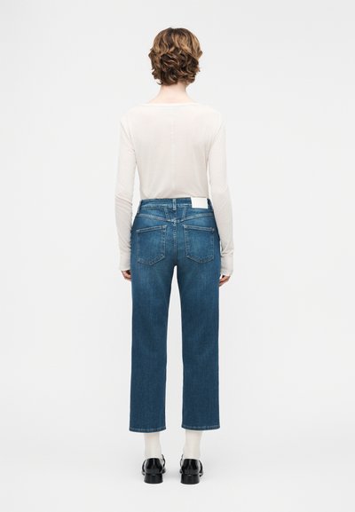 Άτομο με κοντά κυματιστά μαλλιά που φορά λευκό μπλουζάκι με μακρύ μανίκι, μπλε τζιν με ίσια γραμμή, λευκές κάλτσες και μαύρα παπούτσια, στέκεται με την πλάτη προς τα εμπρός.