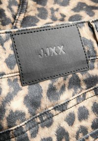 Luipaardprint stof met donkere vlekken op een beige achtergrond, met een zwart leren patch met het etiket "JJXX" in reliëf.