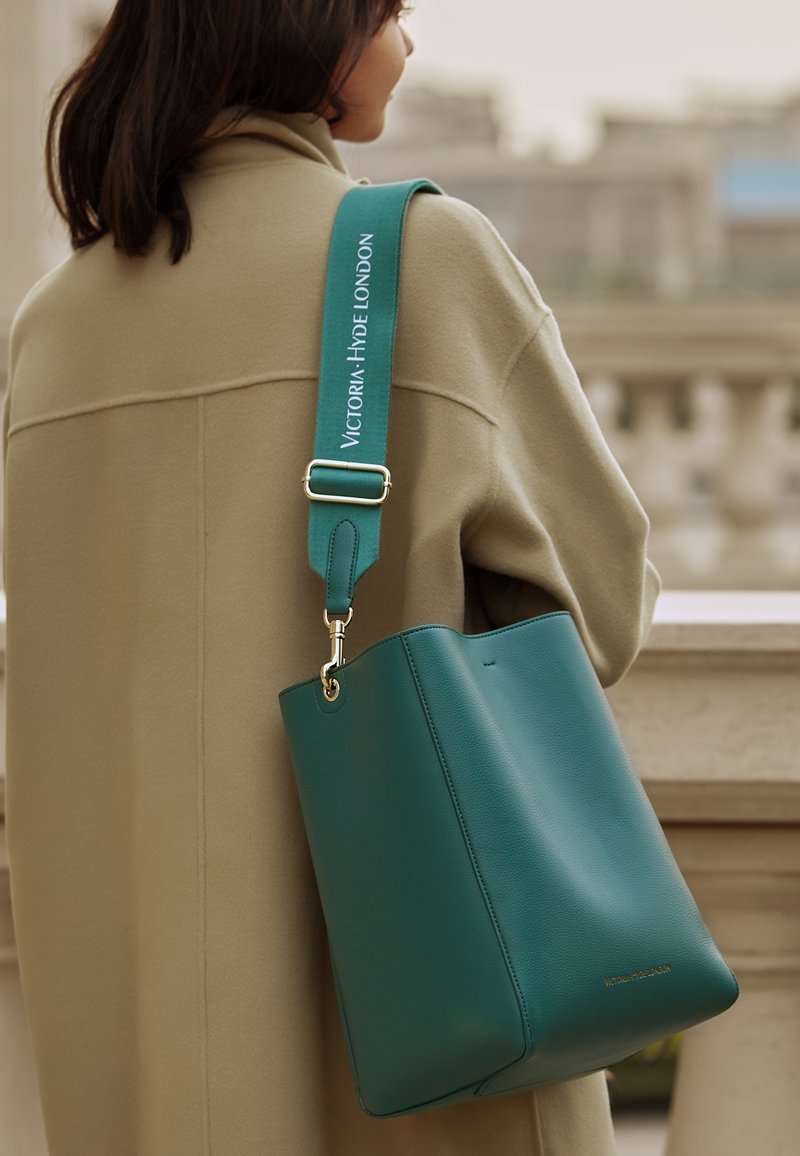 Borsa a secchiello in pelle teal con ampia apertura, dotata di una tracolla in metallo dorato e una tracolla con logo embossato. Texture liscia.
