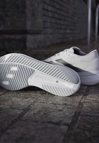 Λευκά παπούτσια τρεξίματος Adidas Adizero σε επιφάνεια από υφή πέτρας, με ένα παπούτσι να δείχνει το σχέδιο της σόλας και το λογότυπο.