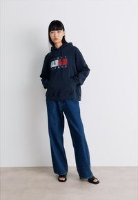 Donna che indossa un hoodie nero di Tommy Jeans, jeans larghi blu e sandali neri con le dita scoperte, in piedi contro un muro bianco con le mani in tasca.