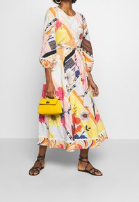 Robe florale colorée avec de longues manches bouffantes, une ceinture à la taille et un ourlet présentant des rayures et des motifs. Accompagnée d'un petit sac à main jaune.