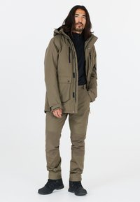 Veste d'extérieur vert olive avec plusieurs poches, zip avant, capuche réglable ; associée à un pantalon assorti et des chaussures noires.