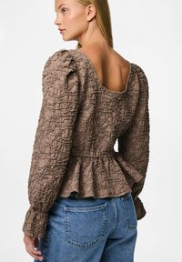 Blusa marrone con texture, scollo a cuore, maniche a sbuffo e orlo peplum, abbinata a jeans in denim blu.