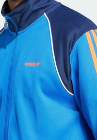 Sininen vetoketjufarkku, jossa tummansinisiä yksityiskohtia, valkoiset koristeviivat ja oranssi Adidas-logo. Valmistettu sileästä, kevyestä kankaasta, jossa sporttinen muotoilu.