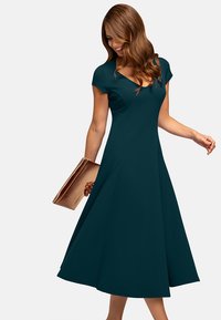 Tassfarbener A-Linien-Kleid mit kurzen Ärmeln und V-Ausschnitt, aus glattem Stoff gefertigt. Der fließende Rock und das taillierte Oberteil betonen die Silhouette. Hält eine goldene Clutch.