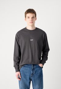 Tricou pe mâneci lungi de culoare gri închis, cu guler rotund, având un mic logo alb pe piept, asortat cu blugi din denim albastru.