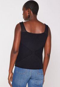 Donna con capelli corti che indossa un top senza maniche nero in maglia e jeans blu, vista di spalle su uno sfondo semplice.