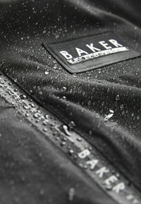 Tissu noir imperméable avec des gouttes d'eau, comportant une étiquette rectangulaire sur laquelle est inscrit "BAKER" en lettres blanches. Surface texturée et finition lisse.
