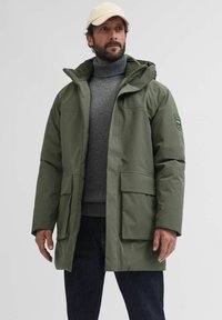 Olivno zelena jakna s podlogo in kapuco, z dvema velikima sprednjima žepoma in zakrpano na rokavu. Uporabljena nad sivo turtleneck puloverjem.