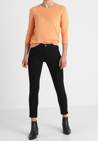 Pull en maille orange clair avec des manches trois-quarts, associé à un jean noir ajusté et des bottines noires. Texture lisse et design classique.