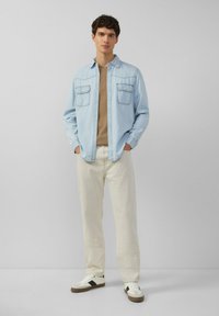 Junger Mann steht mit den Händen in den Taschen, trägt ein hellblaues Denimhemd, ein beiges T-Shirt, off-white Hosen und weiße Turnschuhe.