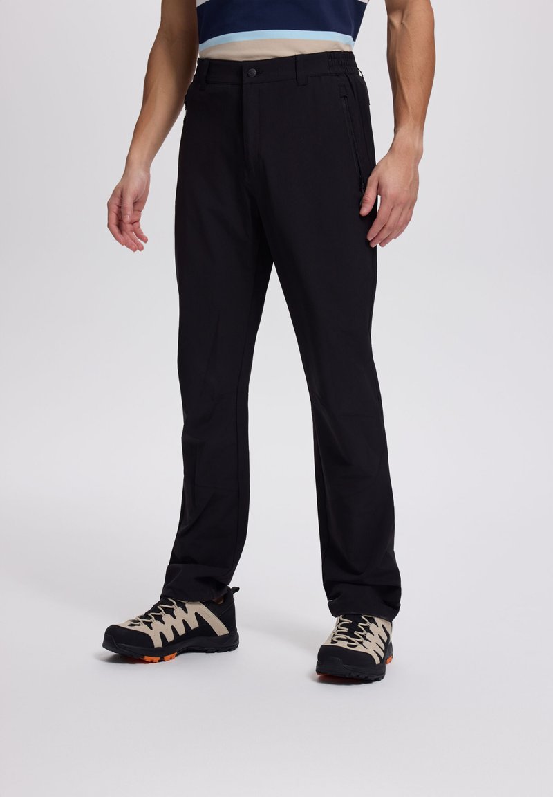Mann trägt schwarze Hose mit geradem Bein, seitlichen Reißverschlusstaschen und beige-schwarze Wanderschuhe vor einfarbigem Hintergrund.