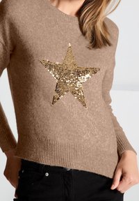 Beige neulepaita, jossa suuri kullanvärinen paljettitähtikuosi edessä, resorit hihoissa ja helmassa. Yhdistetty mustiin farkkuihin.