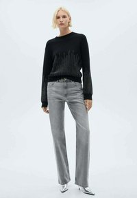 Schwarzer, gestrickter Pullover mit horizontalen Linienakzenten, kombiniert mit hellgrauen, geraden Jeans und silbernen High-Heels, minimalistischem Hintergrund.