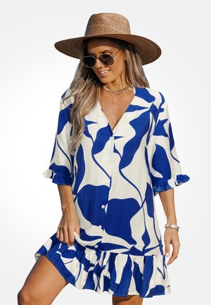 Frau trägt ein blau-weißes, abstrakt gemustertes, knöpfbares Kleid, großen Strohhut, Sonnenbrille, goldene Halskette und Armbänder, lächelt und posiert.