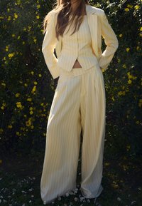 Světle žlutý oblek s tenkými pruhy, obsahující zkrácenou bundu a kalhoty s širokými nohavicemi, s knoflíkovým zapínáním a hladkou texturou. Pozadí ze zeleného listí.