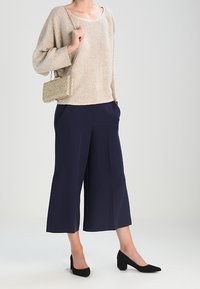 Suéter de punto beige con mangas anchas, culottes navy, zapatos negros de tacón cuadrado y un pequeño bolso dorado texturizado. Diseño simple y moderno.