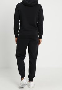 Černá mikina s kapucí a odpovídající zúžené kalhoty, oboje vyrobené z měkké tkaniny. Obsahuje žebrované manžety a elastický pas.