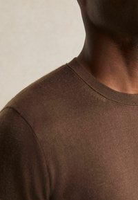 Braunes, tailliertes T-Shirt mit Rundhalsausschnitt, gefertigt aus einem weichen, strukturierten Stoff. Nahtdetails sind entlang der Schulter und des Halsabschlusses sichtbar.