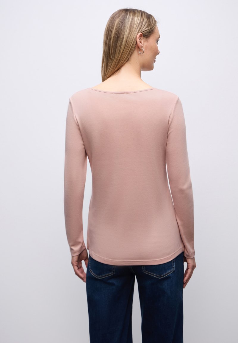 Donna con capelli biondi lisci indossa una maglia a maniche lunghe di colore rosa chiaro e jeans blu scuro, in piedi di spalle contro uno sfondo semplice.