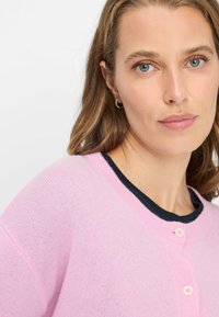 Cardigan rosa con finitura testurizzata, caratterizzato da una scollatura rotonda con bordo blu scuro e due bottoni rosa chiaro sul davanti.