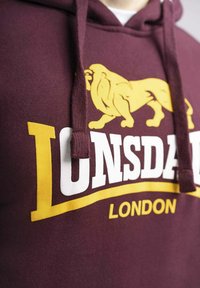 Felpa maroon con cappuccio che presenta una grafica di leone giallo e testo bianco con la scritta "Lonsdale" e "London". Include cordini e una texture di tessuto morbido.