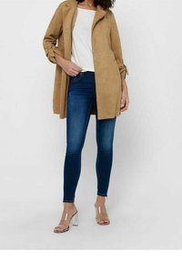 Nainen, jolla on päällä beige polvipituinen takki valkoisen paidan, tummansiniset kireät farkut ja läpinäkyvät korkokengät, seisoo valkoista taustaa vasten.