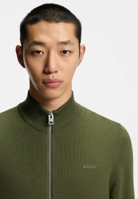 Maglione verde oliva con cerniera e collo alto, con texture a coste. Presenta una zip argentata e un piccolo logo "HUGO" sul lato sinistro.