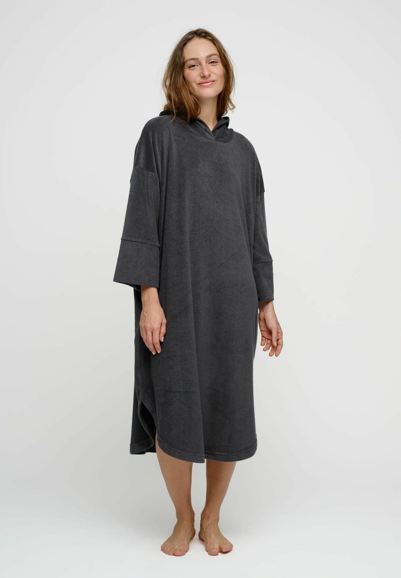 Femme debout pieds nus portant une robe ample à capuche gris foncé, longueur genou, avec des manches trois-quarts, sur un fond blanc uni.