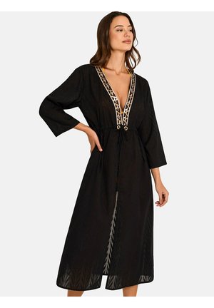 Femme portant une longue robe noire à larges manches, un décolleté en V profond bordé d’un galon à motifs, et une taille à lien coulissant, debout, une main sur la hanche.