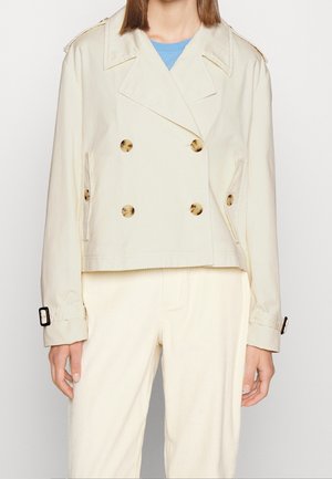 Frau trägt eine kurze beige zweireihige Jacke mit Schildpattknöpfen und geschnürten Manschetten über einer hellen Hose.