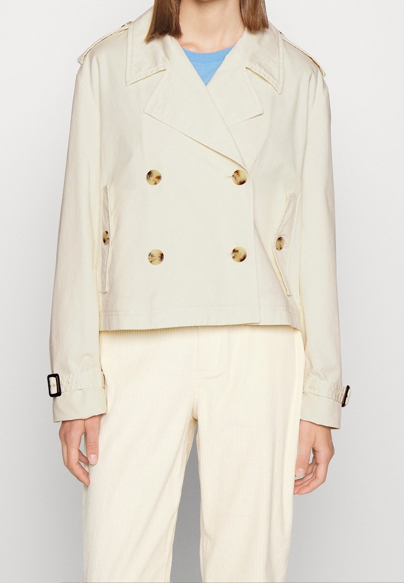 Femme portant une veste courte beige à double boutonnage avec des boutons en écaille et des poignets à boucle, sur un pantalon clair.