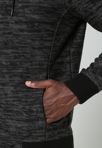 Hand med mörk hudton som vilar i fickan på en mörkgrå texturerad stickad tröja med svarta ribbade manschetter.