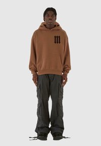Felpa marrone con logo nero sul petto, abbinata a pantaloni cargo neri larghi con tasche laterali e polsini regolabili. Sneakers nere.