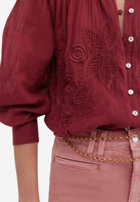 Blouse en coton bordeaux avec des motifs floraux brodés, manches longues, et un devant à boutons, associée à un jean rose clair et une ceinture en chaîne dorée.